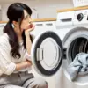日本人女性が洗濯機のふたを開け、洗濯物が汚れたままで落ちていないことにがっかりしている様子。泡立たない洗剤への不満が表れている。
