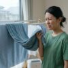 雨の日に部屋干しされた衣類（タオルやシャツ）が、嫌なにおいがして、顔をしかめる表情を浮かべる日本人女性。